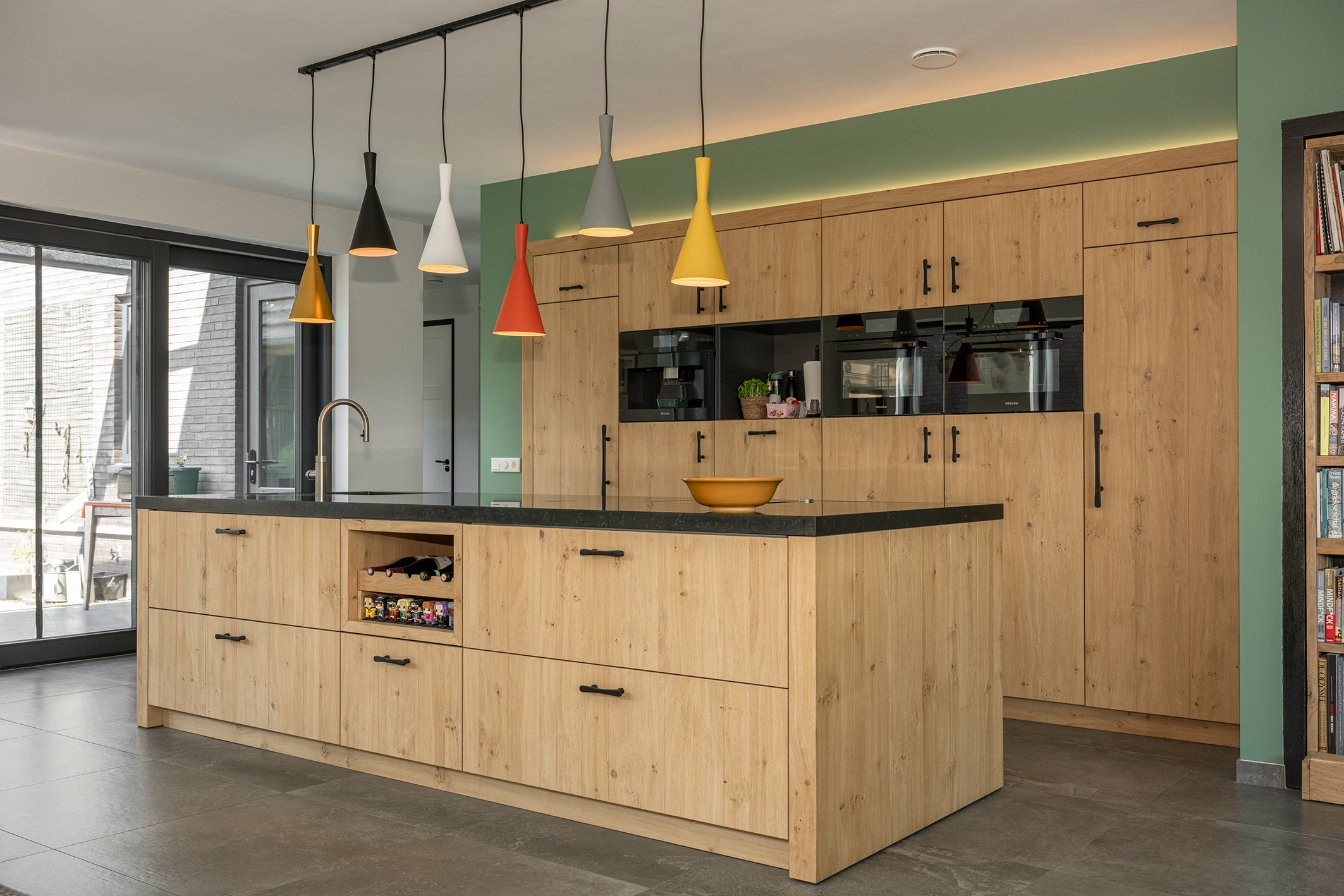 Keuken met houtkleurige kasten, een eiland, donkere werkbladen en hanglampen in verschillende kleuren.