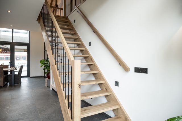 Open houten trap met zwarte spijlen en een houten leuning langs een witte wand.