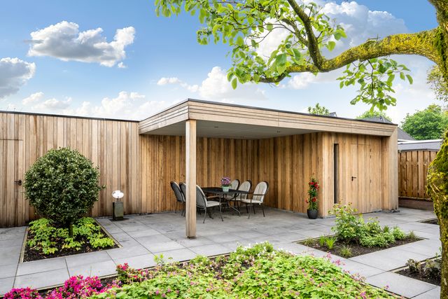 Open overkapping met zitruimte naast een houten gevel en tuin.