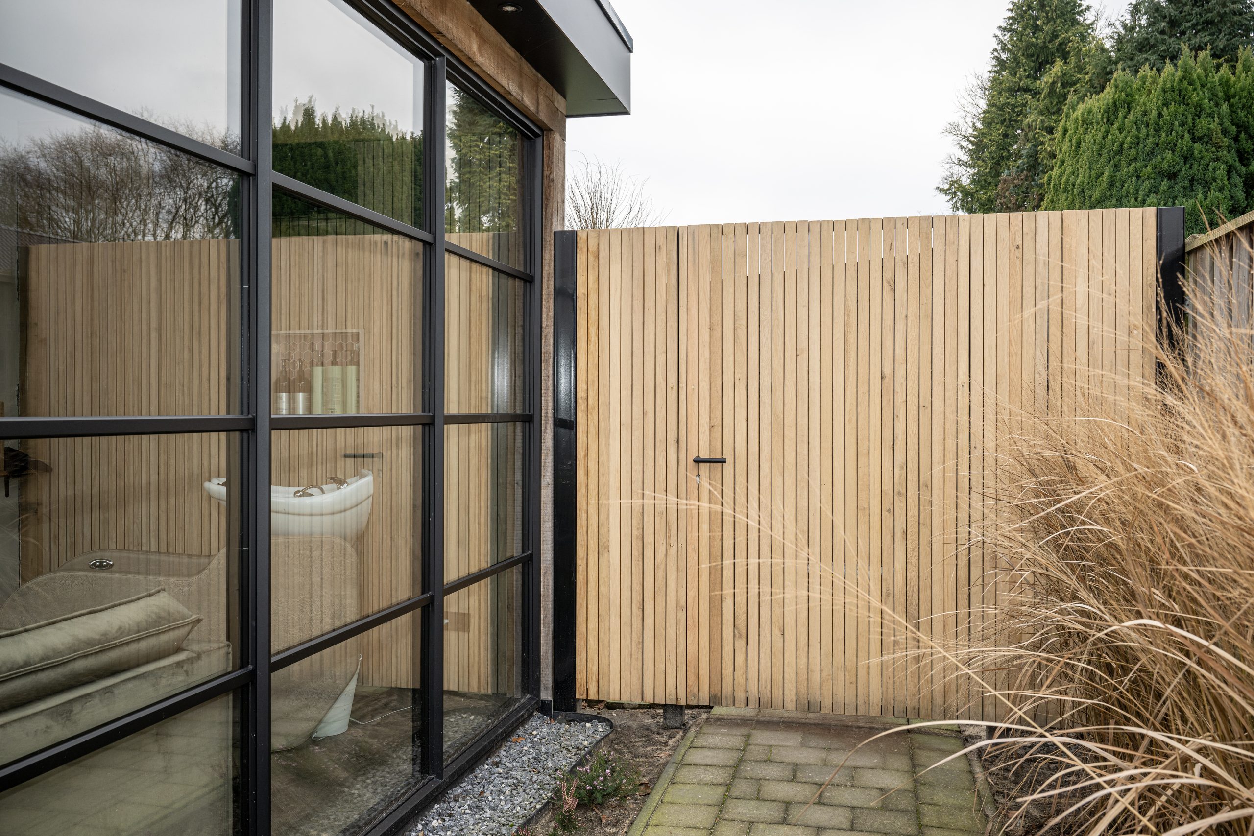 Gesloten houten poort met verticale latten en een donkere deurkruk, naast een glazen wand.