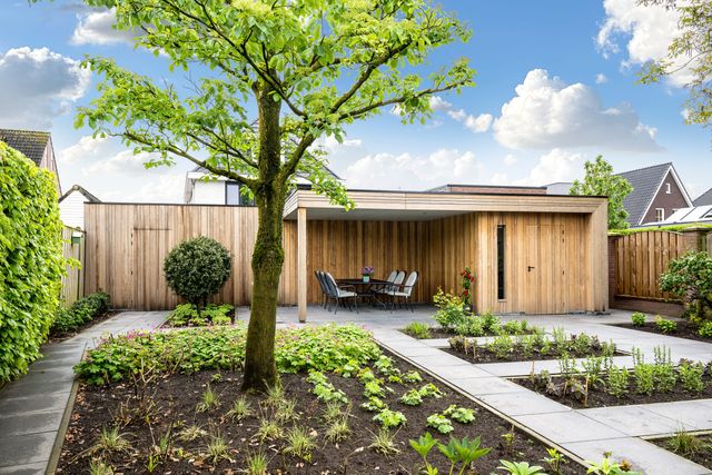 Tuinpad en boom voor een houten buitengebouw met overkapping.