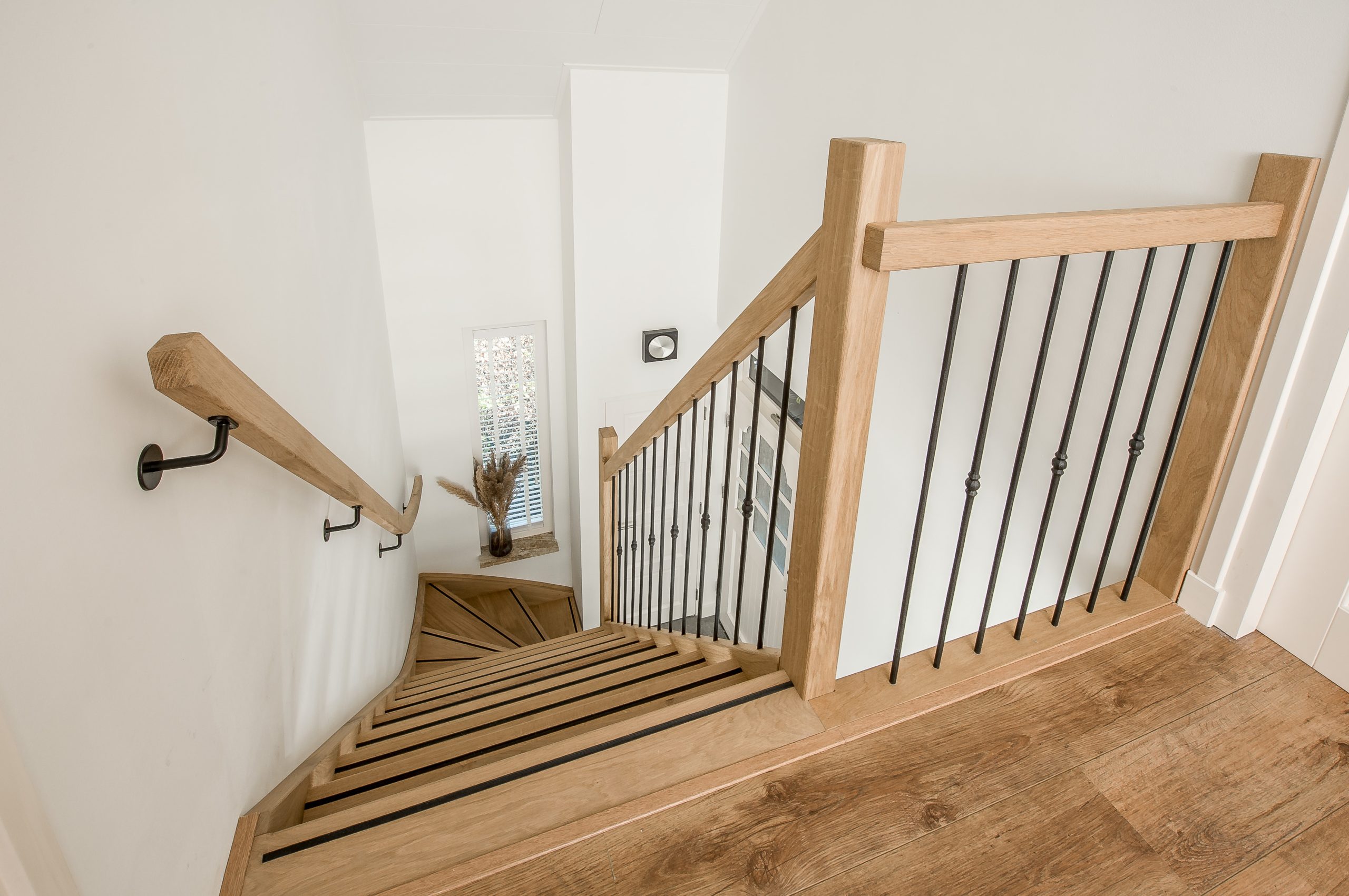 Houten trap met houten leuningen en zwarte spijlen, gezien vanaf de overloop.