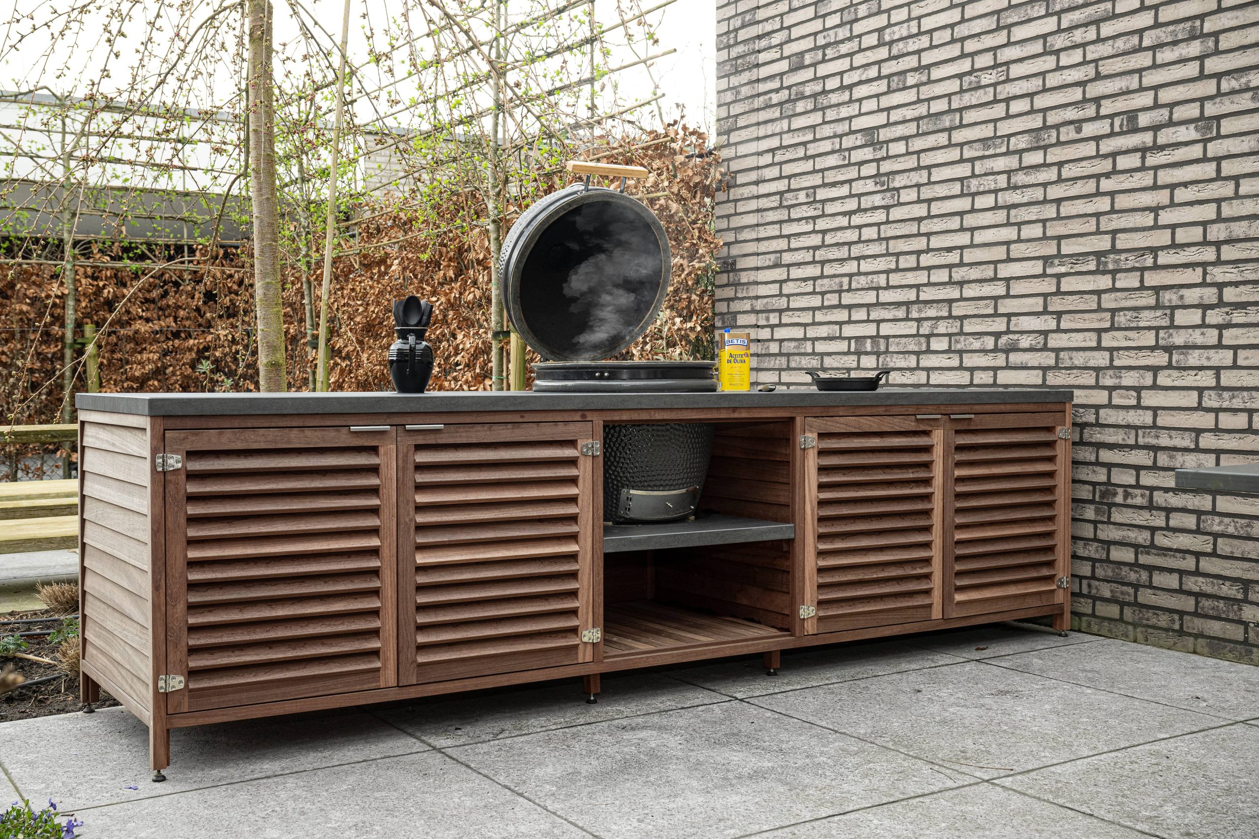 Buitenkeuken met houten kastdeuren, open vakken, een donker werkblad en een ronde barbecue.